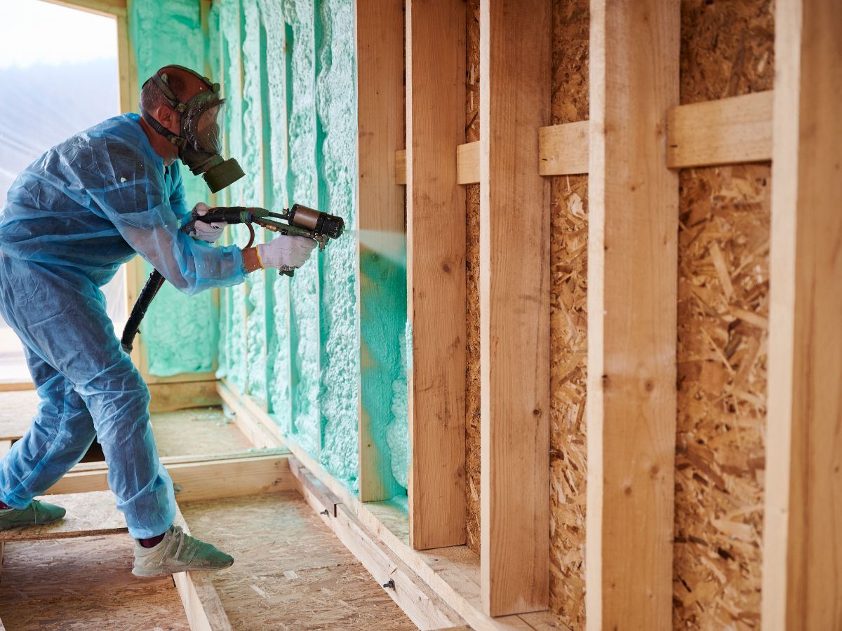 A man in a blue suit applies insulation spray to a wall, demonstrating a construction or renovation process.