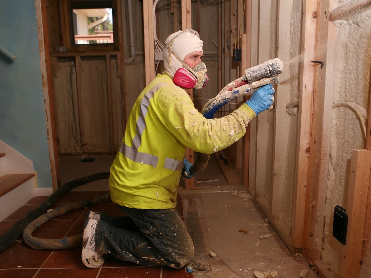 A construction worker in a bright yellow jacket kneels, applying spray insulation to a wall in a partially finished interior space.