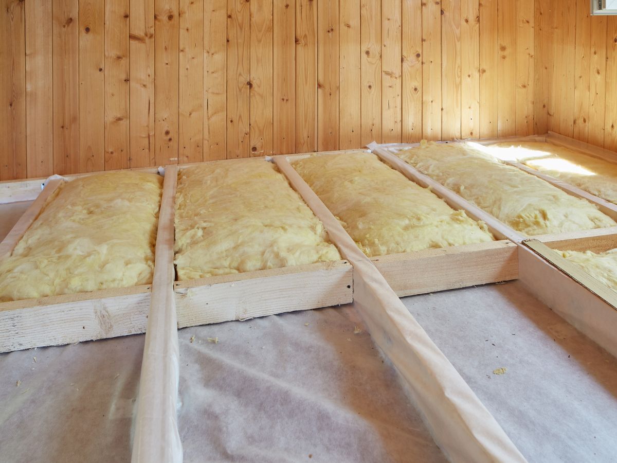 Insulation batts installed in a wooden floor framework, with wooden walls and natural light illuminating the space.
