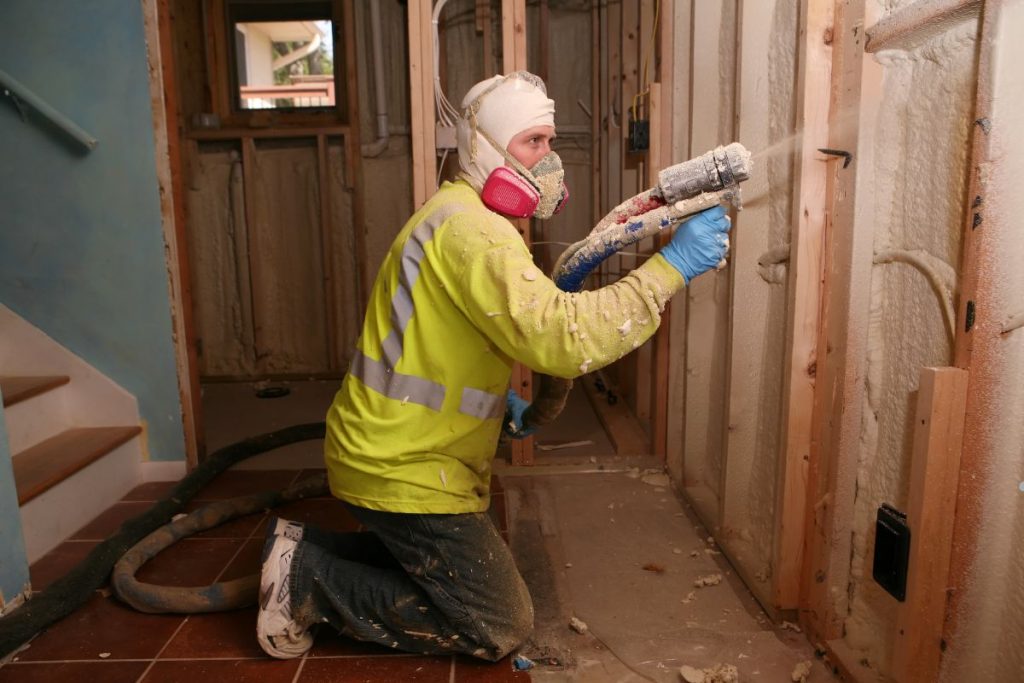 A man in a safety vest and helmet operates a power drill while removing spray foam insulation.