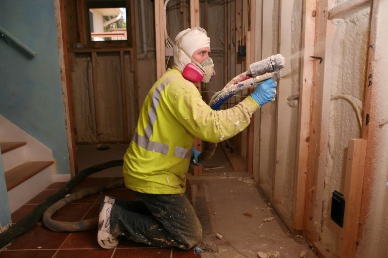 A man in a safety vest and helmet operates a power drill while removing spray foam insulation.